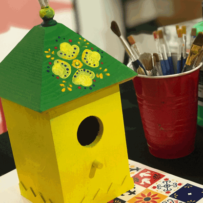Painted green and yellow wooden birdhouse next to a cup with paintbrushed